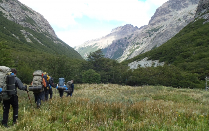 A group of people wearing backpacks hike through a meadow, away from the camera. Tall mountains frame the meadow. 